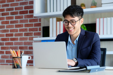 Young asian businessman smiling while working with laptop computer at office, business office lifestyle concept - Powered by Adobe