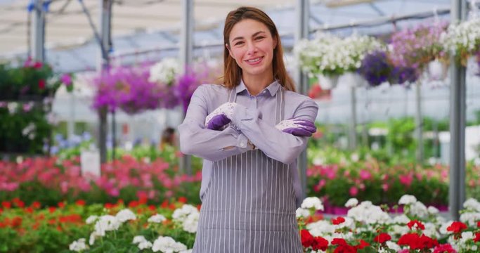 Slow motion of an young woman gardener in apron putting arms crossed satisfied with her work and smiling in camera in a plant shop greenhouse in a sunny day.
