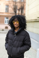 Young african woman with afro hairstyle standing in an urban street. Mixed girl wearing warm jacket on the city background.