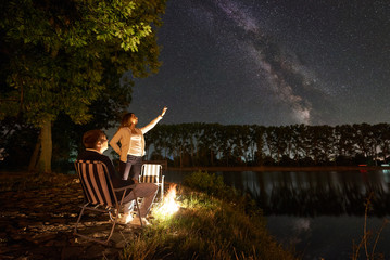 Fototapeta na wymiar Young couple travellers resting near campfire on a lake shore. Man sitting on chair, woman pointing to evening sky full of stars and Milky way above still water and forest on background. Night camping