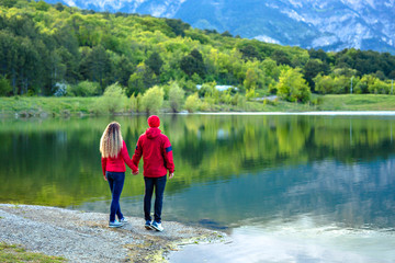 Fototapeta na wymiar A couple in red jackets are standing and looking at the mountains with clouds, symmetrically reflected in the lake. Popular tourist attraction.