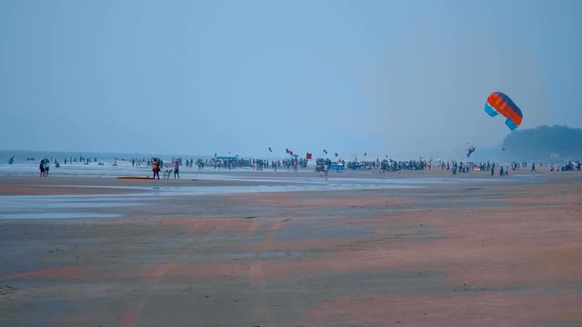 The very long beach of Goa with a huge number of holidaymakers who swim, just walk along the water's edge, and someone rises into the sky on a parasail, towed by a motorboat, India