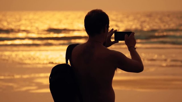 A tourist with a backpack over his shoulder is standing on the shore of the amazing evening sea and shoot a seascape at sunset into his smartphone, Goa, India