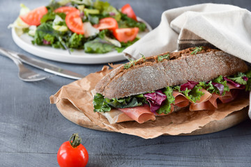 sandwich with baguette, ham, lettuce, kale on gray background, salad from kale and lettuce leaves and tomatoes on a white plate. healthy diet lunch, takeaway sandwich