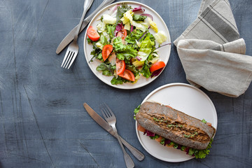 food flat lay sandwich with baguette , ham, lettuce, kale on gray background, salad from kale and lettuce leaves and tomatoes on a white plate.  takeaway sandwich, selective focus