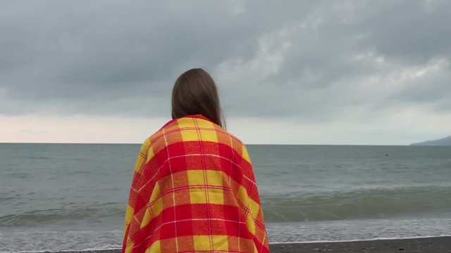 Girl is sitting on the beach wrapped in a bright blanket. Woman looking at the sea having a rest on the seashore