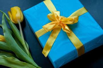 Blue box tied with a festive ribbon and yellow tulips on a black background