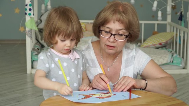 Little cute granddaughter with good looking grandmother sit at children's table and paint goldfish on sheet of paper with colored pencils. Children's creativity. Education concept