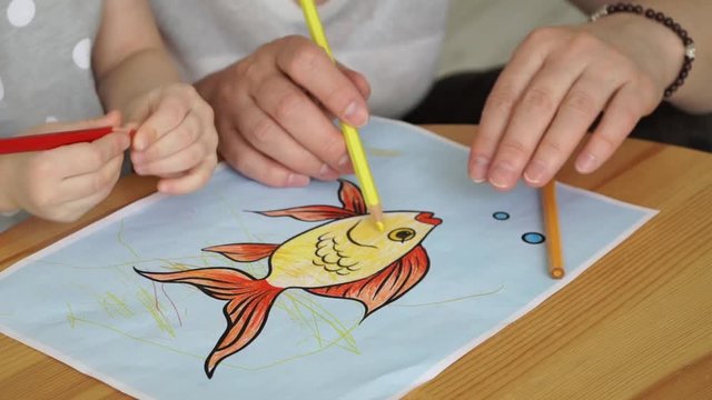 Close up of women's hands and children's hands draw goldfish with colored pencils on sheet of paper. Children's creativity. Education concept