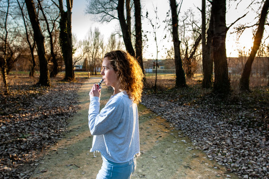 Young woman smoking electronic e-cigarette at the park on sunny day during sunset with smoke.