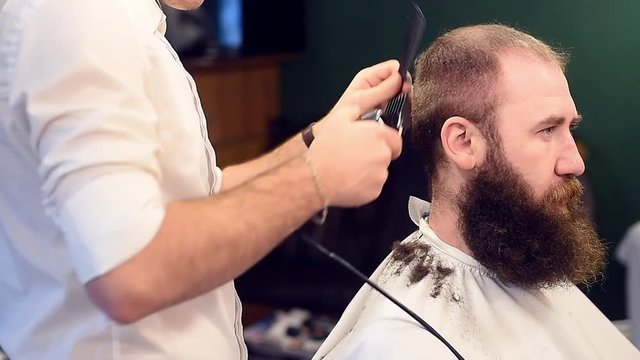 Cropped filming of barber's hands working with electric razor machine, fast moving up and down during cutting hair on head of brunette bearded adult client. Making male haircut process. Side view