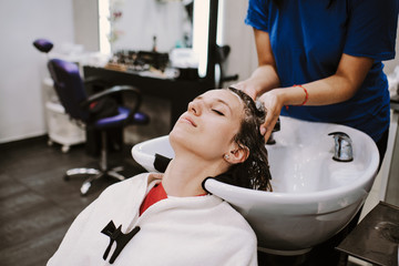 Obraz na płótnie Canvas beauty and people concept - happy young woman with hairdresser washing head at hair salon