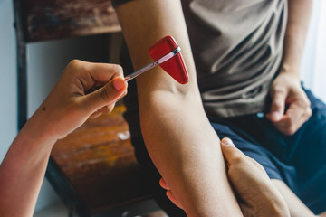 Doctor testing biceps tendon reflex with red hummer jerk on young man arm.