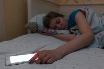 Cute exhausted girl fell asleep with the phone in her hand. Selective focus on phone. White screen. Fatigue, tiredness, overwork. Dependence on social networks, internet addiction, overuse concept.