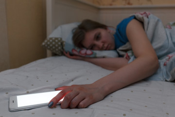 Fototapeta na wymiar Cute exhausted girl woke up in the night. Female can not sleep and takes the phone. Insomnia, nomophobia, sleep disorder. Dependence on social networks, internet addiction, overuse concept.