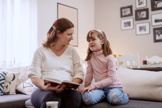 Beautiful happy pregnant mother reading to her lovely daughter fairytale. Living room interior.