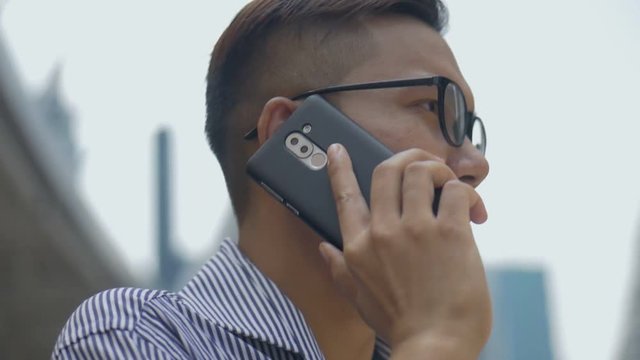 Slow motion portrait asian businessman answers the phone. Asian man with glasses use phone in street near big office building.