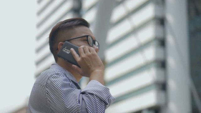 Slow motion portrait asian businessman answers the phone. Asian man with glasses use phone in street near big office building.