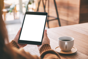 Mockup image of hands holding black tablet pc with blank white screen with coffee cup on wooden table