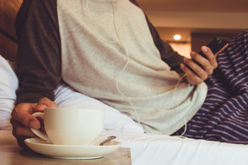 Good morning young man on bed with phone and cup of coffee.