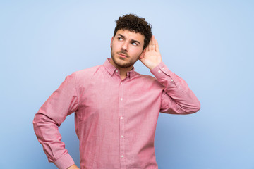 Man with curly hair over isolated blue wall listening to something by putting hand on the ear