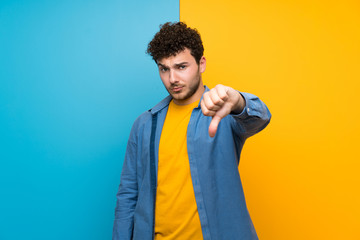 Man with curly hair over colorful wall showing thumb down with negative expression