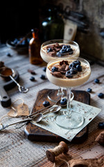 Two champagne glasses with caramel colored panna cotta, jelly, mousse or pudding with chocolate and blueberries on top stands on wooden board