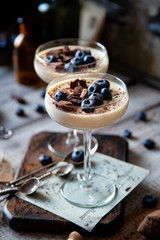 Two champagne glasses with caramel colored panna cotta, jelly, mousse or pudding with chocolate and blueberries on top stands on wooden board