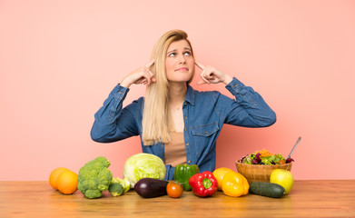 Young blonde woman with many vegetables having doubts and thinking