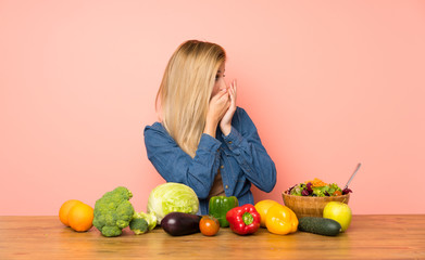 Fototapeta premium Young blonde woman with many vegetables covering mouth and looking to the side