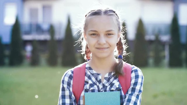 Close up emotional portrait of caucasian smiling teen girl with backpack. Happy cute student teenager with books against the background of house. Childhood and Back to school concept.