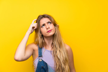 Young blonde woman with overalls over isolated yellow background having doubts and with confuse face expression