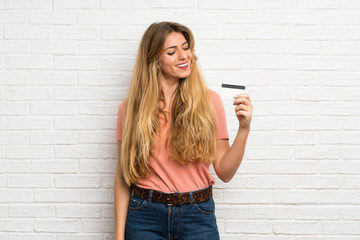 Young blonde woman over white brick wall holding a credit card