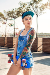 Summertime. Alternative girl skater in cap standing on the city street with penny board looking aside curious