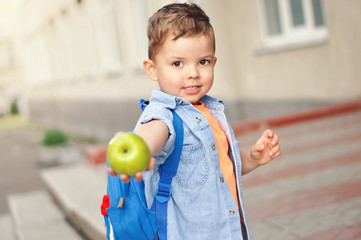 A small three year old preschooler with a backpack on his back shows his green apple for a snack.