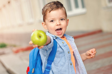 A small three year old preschooler with a backpack on his back shows his green apple for a snack.