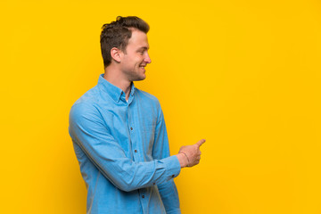 Blonde man over isolated yellow wall pointing back