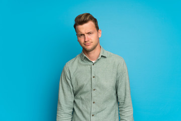 Blonde man over isolated blue wall with sad and depressed expression