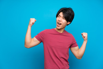 Asian man with red shirt over isolated blue wall celebrating a victory