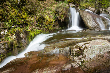 Fototapeta premium Scenic, long exposure, miniature waterfall on a mountain creek during early spring, red, vivid rocks and sunlit, green trees