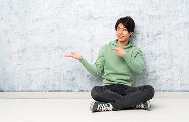 Asian man sitting on the floor holding copyspace imaginary on the palm to insert an ad