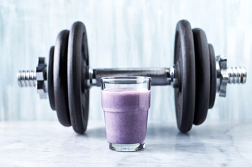 Fototapeta na wymiar Glass of Protein Shake with milk and blueberries and a dumbbell in background. Sports bodybuilding nutrition. Stone / Wooden background. Copy space. 