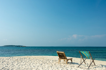 Deckchair and Wooden chairs on the white sandy beach with little waves, blue and bright blue sky.