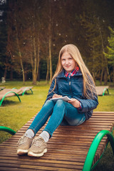 Blonde girl writes a letter sitting on a wooden chaise longue in the Park