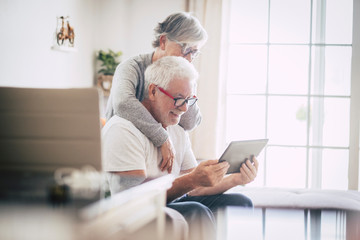 couple of seniors smiling and looking at the same tablet hugged on the sofa - indoor, at home concept - caucasians mature and retired man and woman using technology