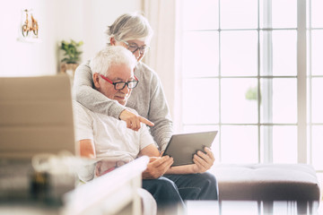 couple of seniors smiling and looking at the tablet - woman hogging at man with love on the sofa -...