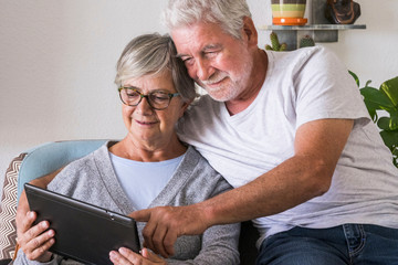 couple of seniors smiling and looking at the same tablet hugged on the sofa - indoor, at home...