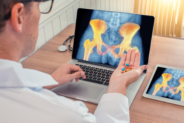 Fototapeta na wymiar Doctor holding pills with x-ray of hips with pain on a laptop. Digital tablet on the wooden desk