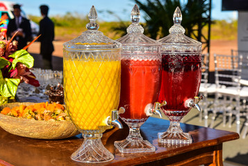 Obraz premium three crystal juice dispensers on wooden table, refreshing colorful fruit drinks for outdoor event, luxury catering service with beverage station, elegant garden party refreshment setup