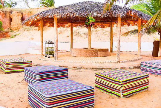 beach lounge area with thatched roof gazebos and colorful ottomans, luxury outdoor event setup on sand, tropical resort relaxation space with decorative string lights, square puffs , piassava kiosk